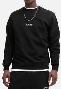 Black sweatshirt featuring a crew neck and ribbed cuffs, with a white logo "JJ Studios" centered on the chest. Smooth fabric texture.