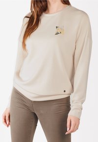 Beige långärmad sweatshirt med en blomsterbroderi-design på bröstet och en liten logotypdetalj vid fållen. Mjuk tyg, avslappnad passform.