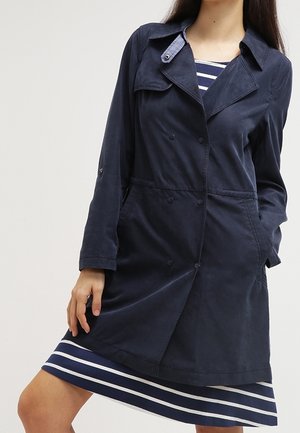 Mujer con un abrigo azul marino de doble botonadura sobre un vestido a rayas azul marino y blanco, de pie con una mano en el bolsillo y la otra al lado.