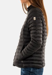 Giacca a vento nera con design trapuntato, caratterizzata da un alto colletto, tasche laterali e un'etichetta con logo sulla manica. Texture liscia.