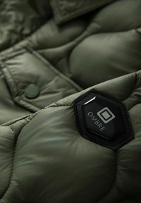 Giacca trapuntata verde scuro con patch esagonale nera del marchio OMBRE e bottoni a pressione visibili su tessuto imbottito.