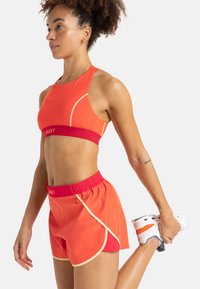 Heldere oranje sportbh met een rode tailleband; heeft gebogen naden. Bijpassende oranje shorts, elastische taille en gele accenten.