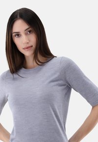 Hellgrauer Kurzarmpullover mit rundem Ausschnitt. Weicher Stoff und dezentes Muster. Das Modell posiert mit Händen in den Hüften vor einem weißen Hintergrund.