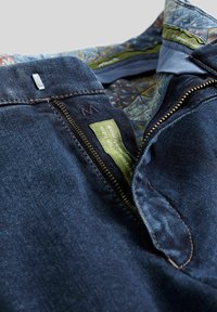 Veste en denim de teinte bleu foncé, avec une fermeture éclair dorée, des coutures audacieuses et une doublure intérieure à motifs. L'étiquette indique "tissu en coton biologique".
