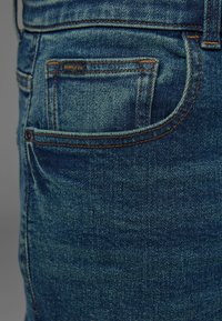Jeans din denim de culoare bleumarin închis, cu material texturat, cusături contrastante și un mic buzunar pe spate cu eticheta "SUPPLY Co."