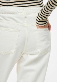 Pantalones de mezclilla de algodón blancos con dos bolsillos traseros, diseño de cintura alta y una textura suave. Parte de una blusa de manga larga a rayas es visible en la manga.