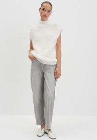 Weißer, flauschiger, ärmelloser Pullover mit einem hohen Kragen, kombiniert mit hellgrauen, lässig geschnittenen Jeans und cremefarbenen Slippern.