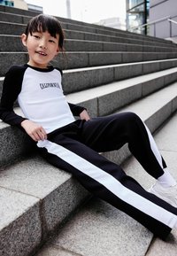 Czarny i biały casualowy strój: longsleeve z napisem "CALIFORNIA", czarne spodnie z białymi paskami po bokach, siedzący na kamiennych schodach.