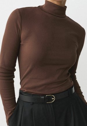 Mujer con un jersey marrón ajustado de cuello alto y acanalado, metido dentro de unos pantalones negros de talle alto con un cinturón negro que tiene una hebilla dorada.