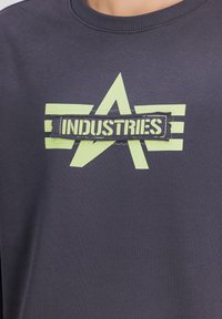 Sötétszürke kapucnis pulóver, középen zöld csillaggal és csíkokkal, a "INDUSTRIES" felirattal egy fekete téglalap alakú részen. Puha tapintás.