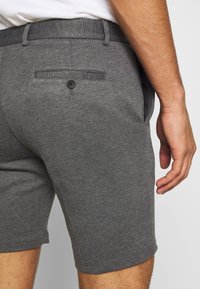 Pantaloni corti grigi realizzati in tessuto testurizzato, con una tasca posteriore con bottone e tasche laterali. La vita è elasticizzata con una finitura liscia.