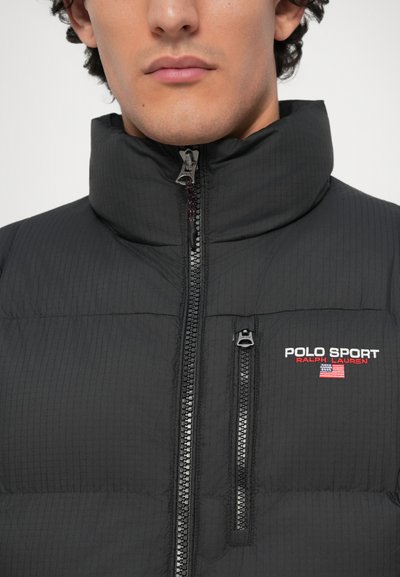 Melnā pūkā jakā ar augstu apkakli, ar priekšējo rāvējslēdzēju un teksturētu audumu. Izšūts "POLO SPORT" logotips baltā un sarkanā krāsā.