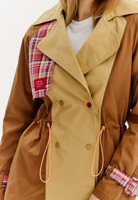 Hnedý a béžový trenčkot s károvaným textilným akcentom. Obsahuje sťahovaciu pásku v páse, dvojité gombíky a nášivku s textom.
