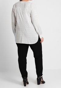 Chemise à manches longues blanche avec un fin motif sombre, présentant un détail de dos yoke ; assortie à un pantalon noir ajusté et des chaussures à hauteur de cheville.