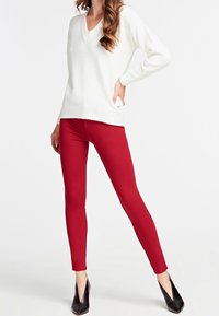 Nainen, jossa on päällään valkoinen V-kaula-aukkoinen neulepaita, punaiset tiukat housut ja mustat teräväkärjiset korot, seisoo valkoista taustaa vasten.