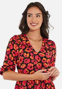 Robe noire avec motif floral rouge et orange, décolleté en V et col volanté. Manches longues avec poignets retroussés. Simples accents de bijoux en or.