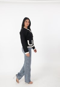 Cárdigan negro de punto con detalles en blanco, que cuenta con botones y un bolsillo frontal. Combinado con jeans rectos de color azul claro y tacones transparentes.
