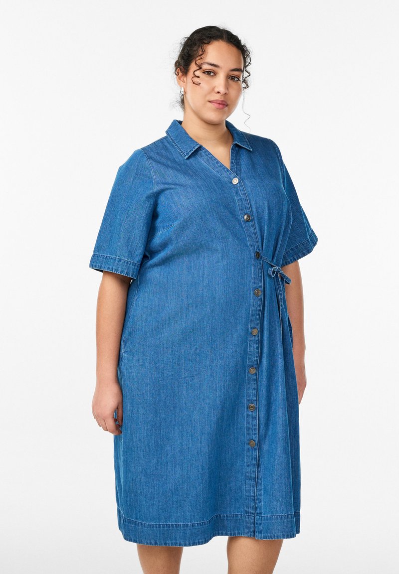 Vrouw met krullend haar die een knielange blauwe denim jurk draagt met korte mouwen, knoopsluiting en een zijstrik, tegen een witte achtergrond.