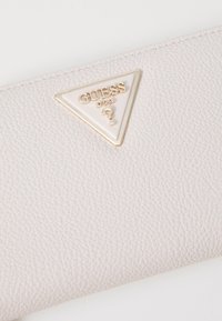 Helles graues Kunstleder-Portemonnaie mit einem dreieckigen weißen und goldenen "GUESS"-Logopatch, strukturiertem Oberflächenmaterial und sauberem Nähtdetail.