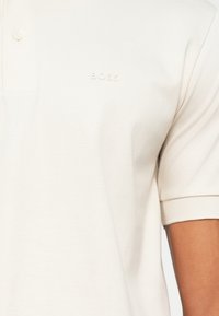 Witte polo-shirt van zachte stof, met een kraag met drie knopen en een subtiel reliëflogo op de borst. Korte mouwen met geribbelde afwerking.