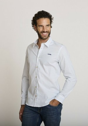 Homme aux cheveux bouclés et barbe souriant, portant une chemise blanche à motifs boutonnée et un jean bleu, debout avec une main dans la poche.