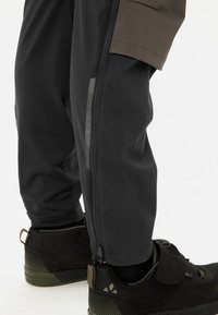 Zwarte sportbroek met een ritsfunctie bij de enkel, gecombineerd met donkere schoenen met een gestructureerd oppervlak en groene accenten.