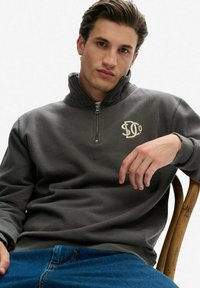 Grijze kwart-rits pullover met een geribde kraag en geborduurd logo op de linkerborst. Zacht materiaal, relaxte pasvorm, gecombineerd met blauwe jeans.