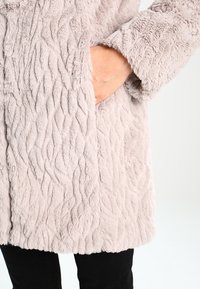 Main dans la poche d'un manteau en fausse fourrure beige clair texturé porté sur un pantalon noir devant un fond uni.