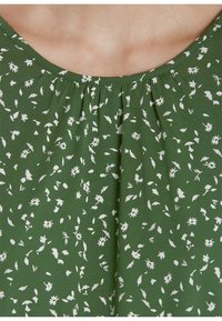 Tissu vert à petit motif floral blanc avec des plis froncés sous un col rond sur un vêtement.