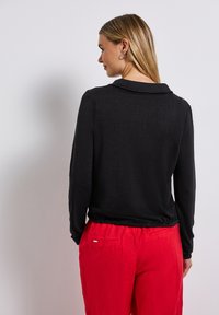 Černý top s dlouhým rukávem a límečkem, hladká textura, přiléhavý silueta, v kombinaci s volnými červenými kalhotami s zadní kapsou.