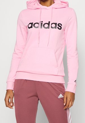 Pink hoodie with a front pocket, featuring black "adidas" logo, drawstrings, and ribbed cuffs. Paired with brown pants and white stripes.