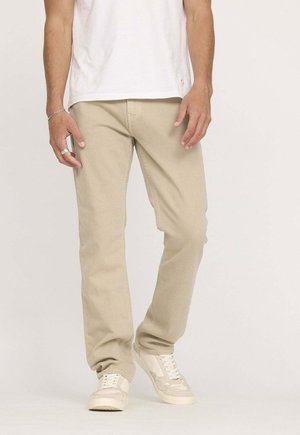 Pantalon en coton beige à coupe droite, poches avant, texture légère, porté avec des baskets blanches et un t-shirt blanc.