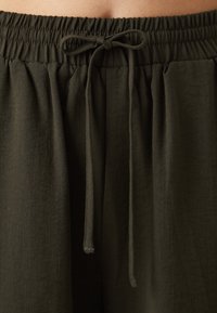 Mørkegrønt, lett stoff med elastisk midjebånd og snøring. Teksturen virker myk og lett krøllete.