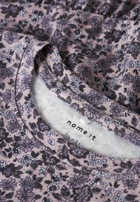 Nahaufnahme eines gefalteten violetten Stoffes mit kleinem dunklem Blumenmuster und einem Halsausschnitt-Etikett mit der Aufschrift "name it" und Größe 1½ - 2 Jahre.