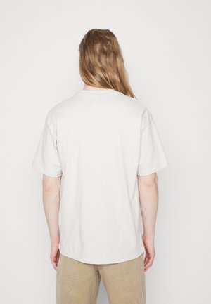 Une personne avec de longs cheveux châtain clair se tient face à un mur blanc, vêtue d'un t-shirt blanc à manches courtes et d'un pantalon beige.