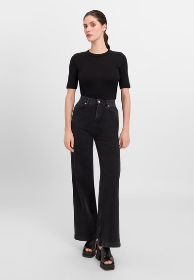 Zwarte high-waisted jeans met wijde pijpen, denim stof, twee voorzakken en details met zilveren knopen. Gecombineerd met een nauwsluitend zwart T-shirt met korte mouwen.