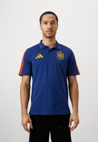 Jūras zila polo krekls ar sarkaniem un dzelteniem svītrainiem akcentiem. Ieskaitīts izšūts Spānijas logo un Adidas zīmols dzeltenā krāsā uz krūtīm.