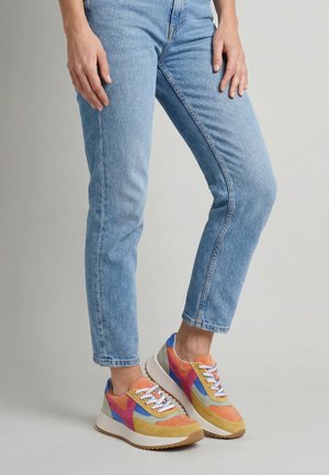 Person trägt hellblaue Jeans und bunte Sneakers mit orangenen, pinken, blauen und gelben Akzenten und steht auf grauem Boden.