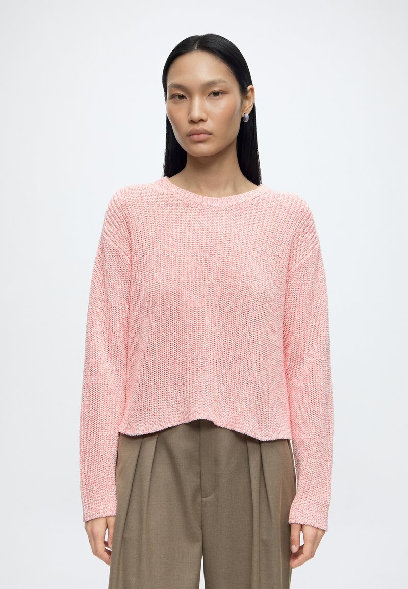 Femme aux cheveux noirs raides portant des boucles d'oreilles argentées, un pull en tricot rose et un pantalon plissé marron taille haute sur un fond uni.
