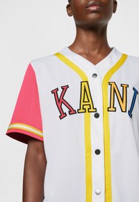 Valge pluus lühikeste roosade varrukatega, millel on kollased ja mustad äärised ning mitmevärvilised tikitud tähed "KANI". Hõbedased nupud.