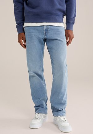 Homme portant un jean droit bleu clair, des baskets blanches et un sweat-shirt bleu marine, debout sur un fond uni.