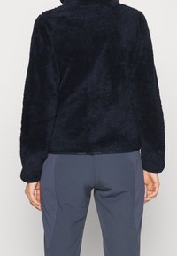 Pull en polaire bleu marine avec un col montant, surface texturée et manches longues, associé à un pantalon gris. Mettez l'accent sur le confort et la chaleur.