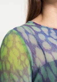 Durchsichtige Bluse mit einer Mischung aus grünen und blauen Mustern, rundem Ausschnitt und strukturiertem Stoff, der leicht wirkt.