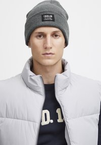 Gray knit beanie with a black label, gray padded vest with high collar, and dark blue sweatshirt featuring a stitched light beige letter design.