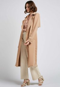 Manteau trench beige avec une texture lisse, devant ouvert et grands revers, superposé à un chemisier à motifs et un pantalon large crème. Sandales à talons.