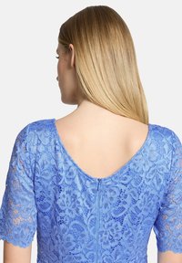 Femme blonde aux cheveux raides portant une robe en dentelle florale bleue à manches courtes et dos en décolleté plongeant, vue de dos.