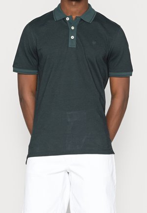 Ciemnozielona koszulka polo z fakturowanej tkaniny, z zapięciem na trzy guziki oraz kontrastującym kołnierzykiem i mankietami. Białe szorty widoczne poniżej.