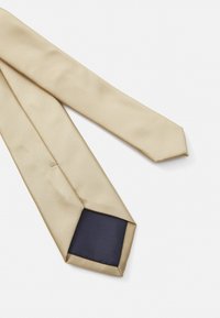 Cravate beige avec une finition texturée, présentant un dessous en soie bleu foncé. La cravate a une forme traditionnelle étroite et une pointe pointue.