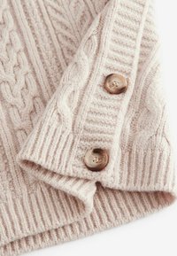 Knitwear-kaapu pehmeas bežis, millel on soonikstruktuur, lõppenud servad ja kaks ümmargust, pruuni tooni nuppu. Hubane, soe disain üksikasjaliku õmblemisega.