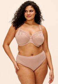 Krølhåret kvinde iført beige bh med blondedetaljer og matchende højtaljede trusser står mod en neutral baggrund og kigger til siden.